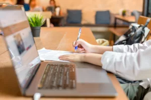 Une personne qui rempli un document papier avec son ordinateur portable allumé posé sur la table