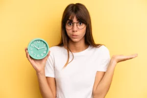 Jeune femme qui n'arrive pas à lire l'heure sur son horloge en anglais devant un fond uni jaune pâle.