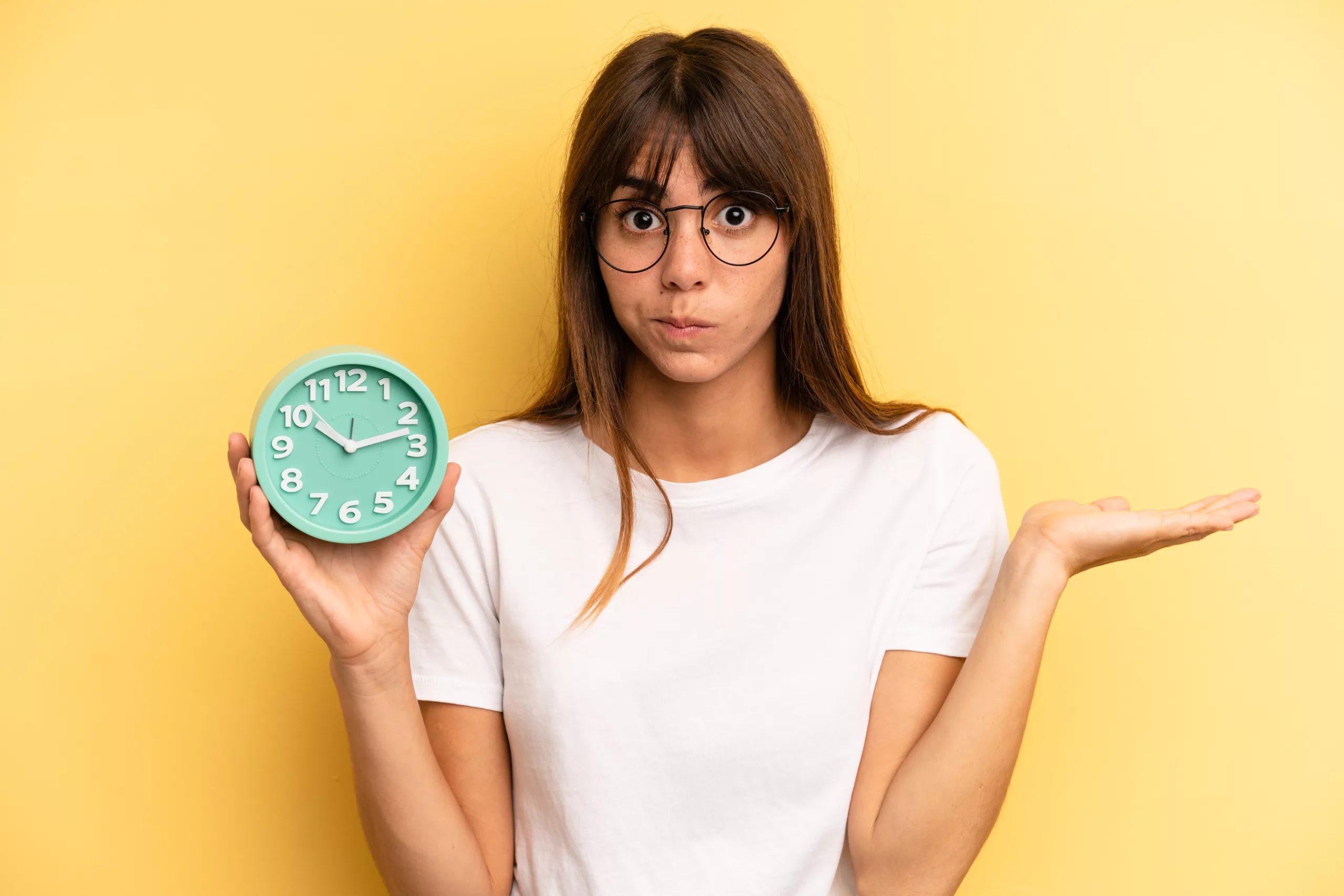 Jeune femme qui n'arrive pas à lire l'heure sur son horloge en anglais devant un fond uni jaune pâle.
